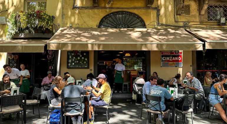 Travelers dine at outdoor tables in front of a cafe with a Cinzano sign and a waiter standing in the doorway.