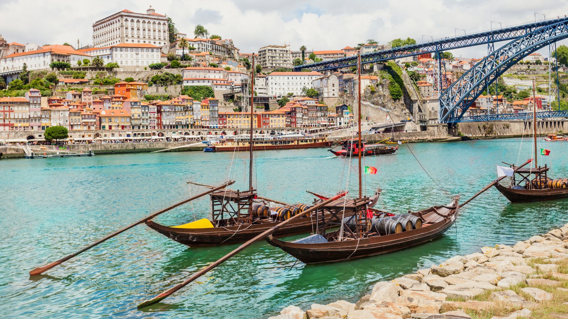 Traditional wooden boats carrying barrels float on the Douro River in Porto, offering a classic travel view of the city skyline and Dom Lu&iacute;s I Bridge in the background.