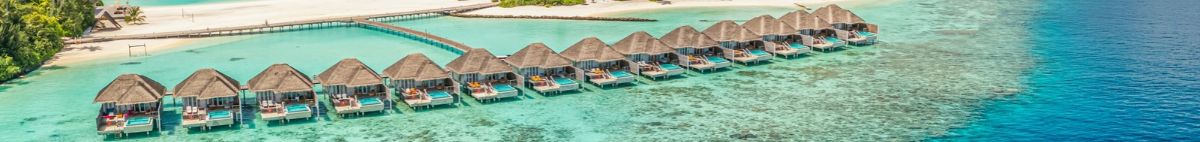A row of overwater bungalows for travel extending into the turquoise ocean from a sandy beach.