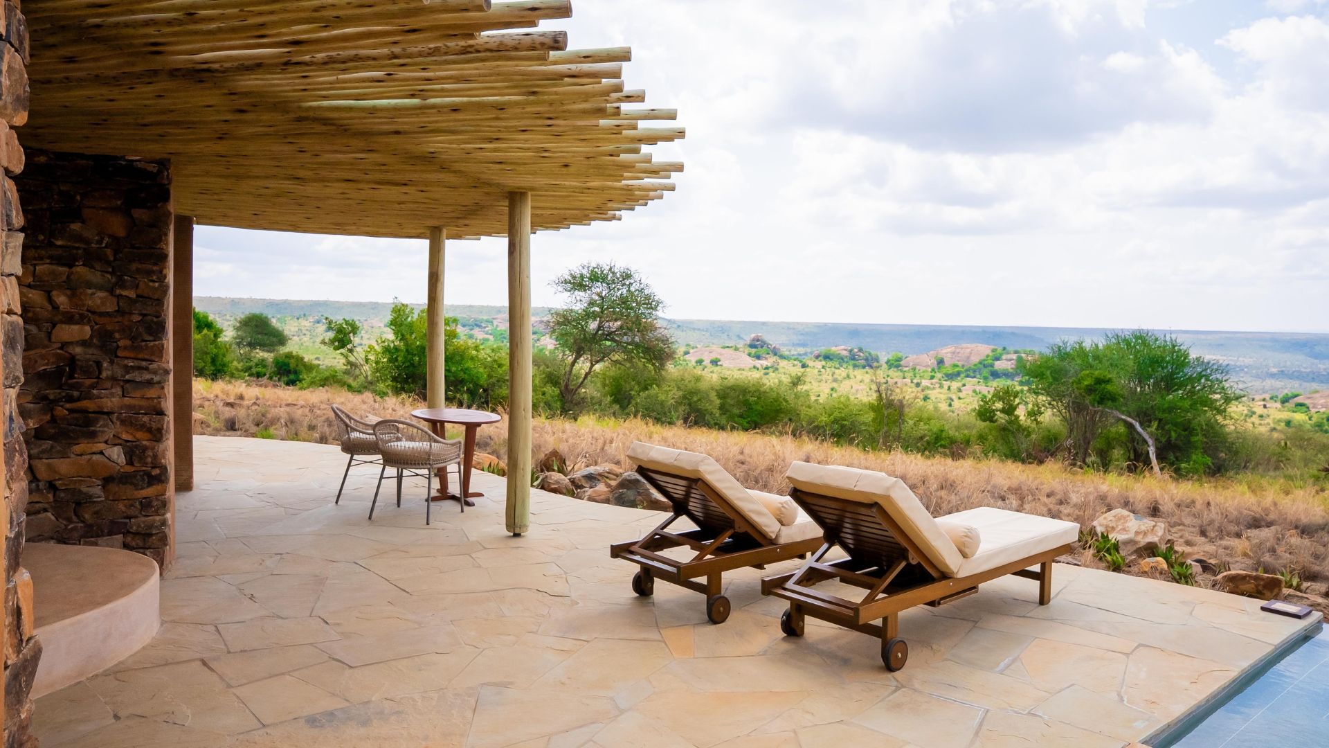 A stone patio with two lounge chairs under a pergola and a small table set for travel, overlooking a savanna landscape.