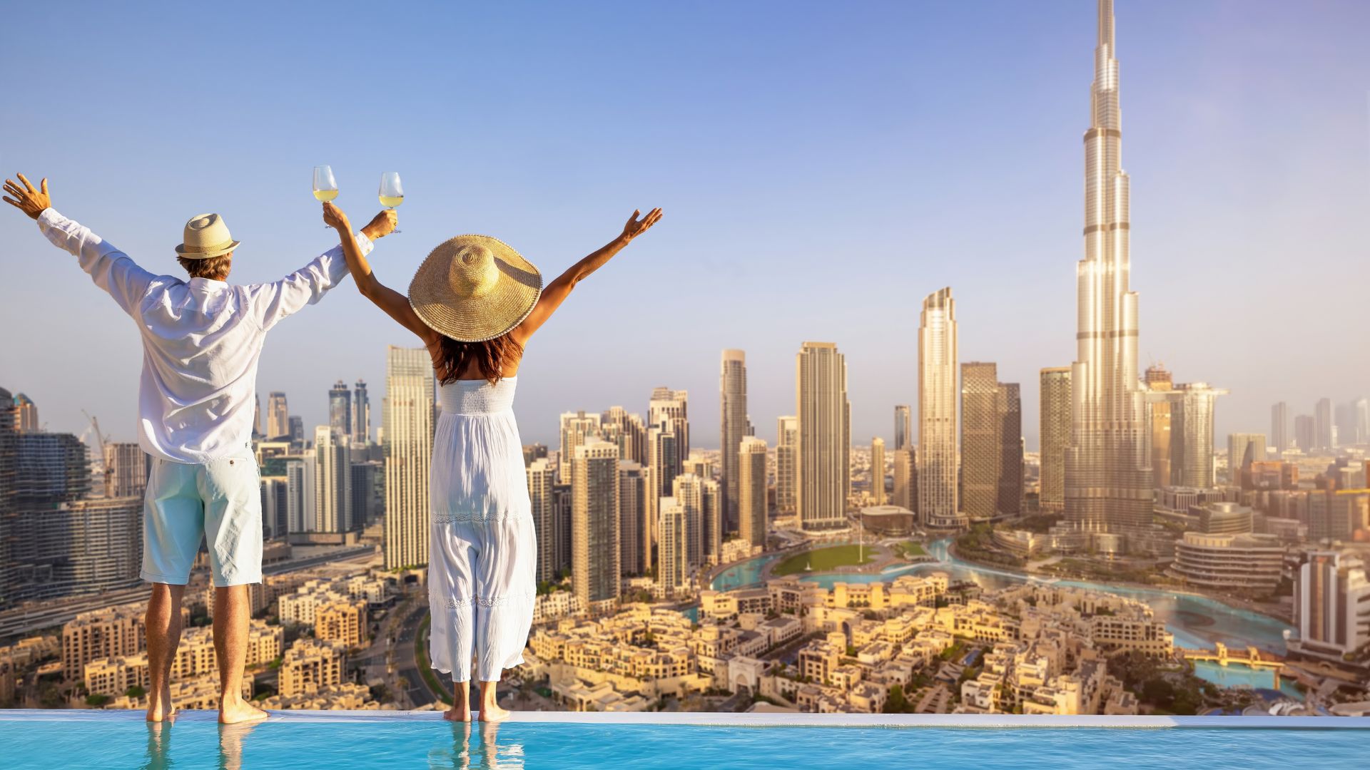 A couple celebrates during their travels, arms raised with wine glasses at the edge of an infinity pool overlooking the Dubai skyline and the Burj Khalifa.