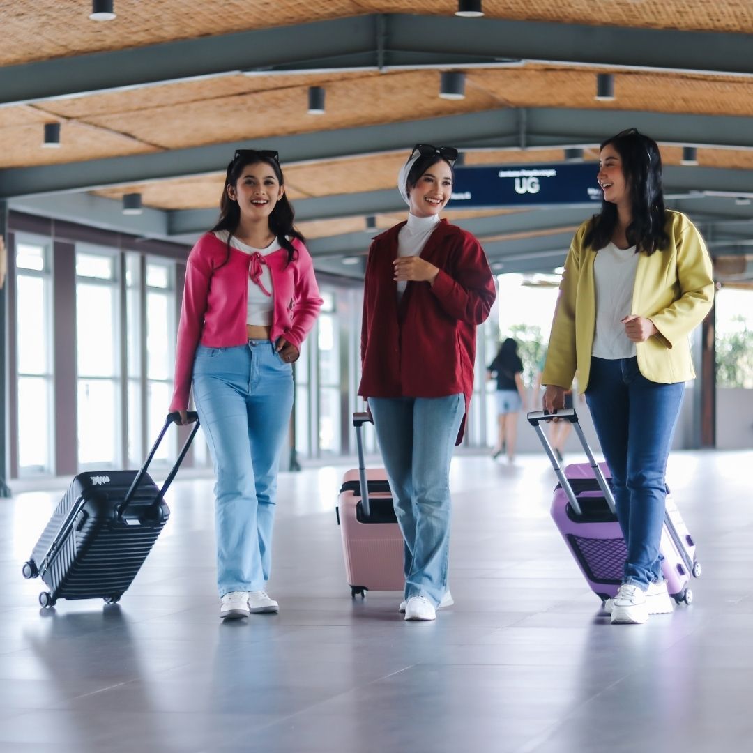 Three women smile while walking through a modern building concourse, each pulling a piece of travel luggage.