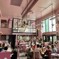 A lively pink-themed café filled with people sitting at pink chairs and tables, surrounded by floral decorations, hanging plants, and large windows letting in natural light.