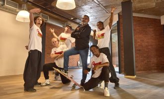 Six men pose indoors; five wear matching white shirts with a red and yellow logo, one stands in the center wearing a black jacket and glasses. Some crouch or kneel, one leans on a skateboard, all appear cheerful and playful.