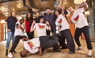 A group of people pose cheerfully indoors. Four are wearing HOT 102.7FM t-shirts, with two breakdancing in front. Others stand behind them, some smiling and making peace signs, in a brightly lit modern office space.