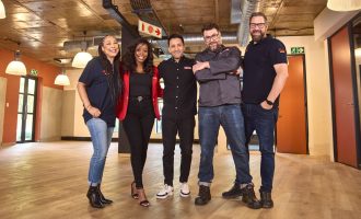 Five people stand smiling together in a spacious, modern room with wooden floors and industrial ceilings. Three men and two women, all casually dressed, pose with arms around each other, conveying a friendly atmosphere.