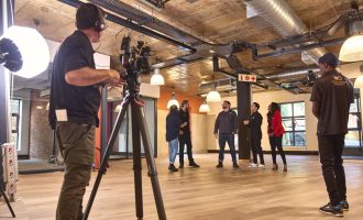 A cameraman films four people standing and talking in a modern, industrial-style room with exposed pipes and ceiling lights. Another crew member stands to the side, facing the group.
