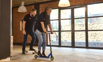 Two people in casual clothes ride a skateboard and a scooter indoors on a wooden floor, smiling and enjoying themselves near large windows and brick walls.