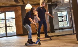 Two people ride in an office; one rides a blue scooter and the other rides a skateboard. Both wear black shirts and jeans, smiling as they move on a wooden floor with brick walls and large windows in the background.