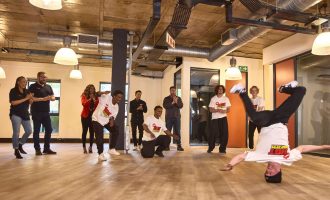 A group of people watch and applaud as dancers in white t-shirts perform breakdancing moves in a spacious room with wooden floors and industrial lighting. One dancer is in a headstand, while another is mid-spin.