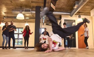 A man breakdances on the floor in an office space while others in casual and business attire stand around watching and cheering him on. The room has a wooden floor, exposed ceiling pipes, and large windows.