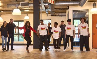 A woman in a red jacket leads a group dance practice in a spacious room. Several people, some in matching “Gritt” t-shirts, watch and prepare to join in. Large windows and hanging lights are visible.