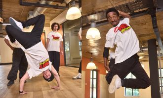 Two men perform breakdancing moves in a studio, wearing white t-shirts and black pants; one does a handstand freeze while the other jumps mid-air. Two people watch in the background.
