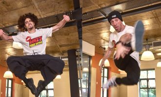 Two men, both wearing white t-shirts and black pants, are captured mid-air performing energetic dance or acrobatic moves inside a modern room with large windows and exposed ceilings.