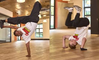 Two breakdancers perform moves indoors; one balances on one hand with legs in the air, while the other does a headstand with bent knees. Both wear white T-shirts, black pants, and sneakers in a spacious room with wooden floors.