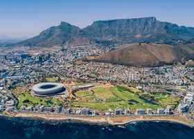 Cape Town. Where city, ocean and mountains meet. Aerial view of Cape Town, South Africa, featuring the coastline, Cape Town Stadium, and the city with Table Mountain and Lions Head in the background under a clear blue sky.