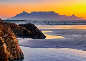 Table Mountain glowing in the soft light of a Cape Town sunset. Rocky beach at sunset with Table Mountain in the background, calm waves, and two people sitting on rocks observing the colorful sky reflected in tide pools.