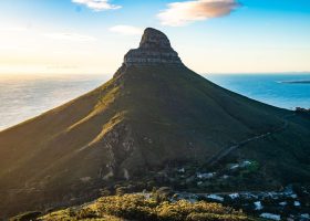 From the summit of Lion’s Head, Cape Town unfolds along the Atlantic coast. A prominent, cone-shaped mountain rises beside the ocean at sunset, with a winding road leading up and scattered houses at its base. The sky is clear with a few clouds and sunlight illuminating the landscape.