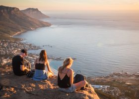 Early hikers climb Lion’s Head to watch the city wake beneath them. Three people sit on a rocky ledge overlooking a coastal landscape at sunset, with mountains, a bay, and a town below. The sky is clear, and the ocean stretches out to the horizon.