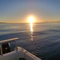 A bright sunrise over calm, blue ocean water with distant mountains on the horizon; part of a small boat is visible in the foreground.