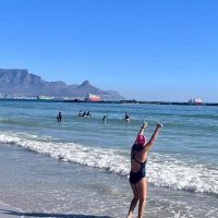 A person in a blue swimsuit and red cap stands on a sandy beach with raised arms, facing the ocean. Several people are in the water, and distant mountains are visible under a clear blue sky.
