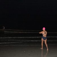 A person wearing a pink swim cap and dark swimsuit stands on a beach at night, facing the ocean with one arm extended forward. The sea and sky are dark, with only a few faint lights visible in the distance.