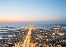 Aerial view of the Palm Jumeirah in Dubai at dusk, a popular travel destination featuring the illuminated palm-shaped archipelago and its central road extending into the Persian Gulf.