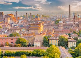 Aerial view of the historic city of Bologna, Italy, featuring its terracotta rooftops, domed churches, and the Asinelli Tower rising above the skyline.